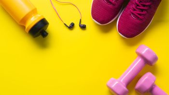Flat lay of red sport shoes, pink dumbbells, earphones, bottle of water on yellow background