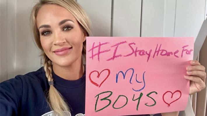 Carrie Underwood holding a pink sign that says "#IStayHomeFor My Boys" with two hearts