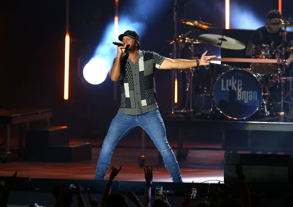 Luke Bryan on stage singing into a microphone while dancing wearing a patterned shirt and skinny jeans