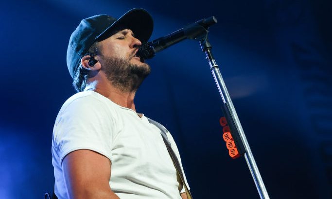 Luke Bryan on stage with white t-shirt and blue jeans on and a black baseball hat with a yellow guitar around his neck singing in to a microphone stand