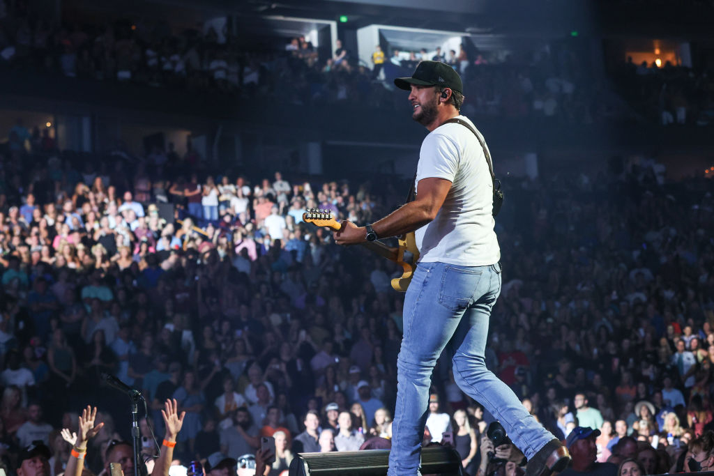 Luke Bryan on stage in white shirt and blue skinny jeans with a baseball hat on singing and playing guitar