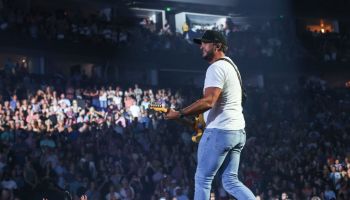 Luke Bryan on stage in white shirt and blue skinny jeans with a baseball hat on singing and playing guitar