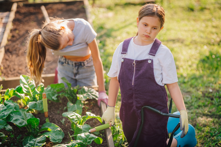 Young girls gardening together, one looking bored while the other is busy