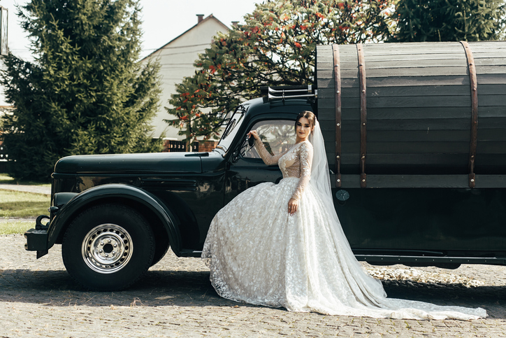 Woman in wedding dress poses beside a vintage truck in a garden during daytime celebration in an outdoor setting with trees and flowers around