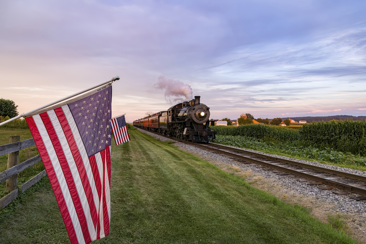 A View of a Classic Steam Passenger Train Approaching, With American Flags Attached to a Fence on a Sunny Summer Day
