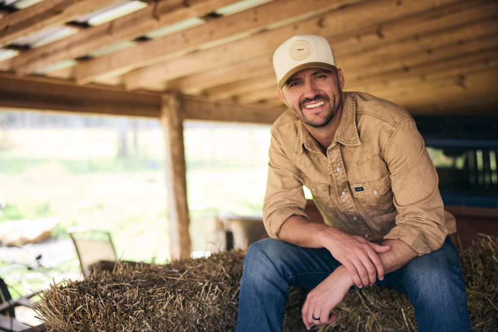 A smiling man wearing a tan jacket and baseball cap sits on a pile of hay in a wooden structure.