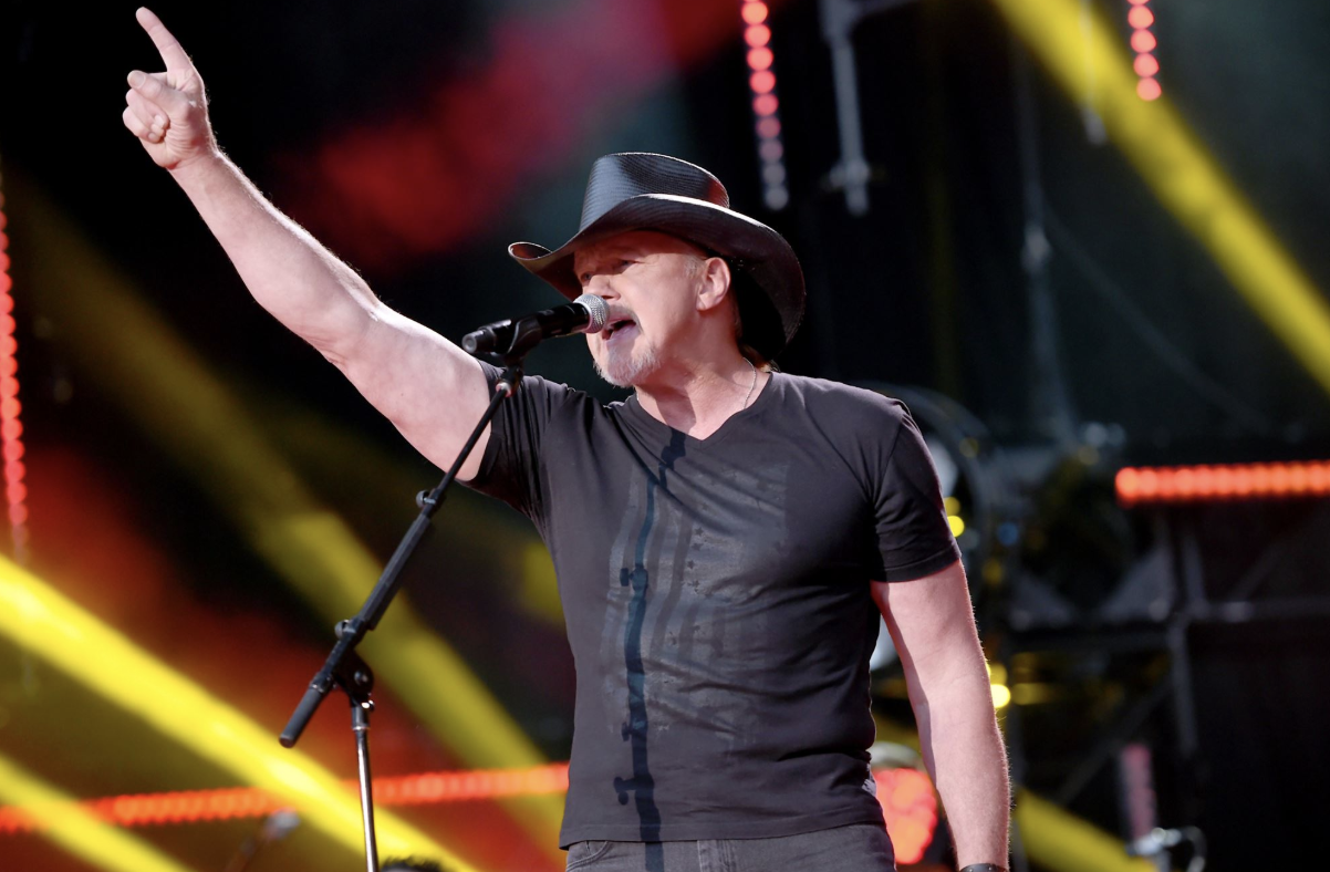 A man wearing a cowboy hat and t-shirt performing on stage, with his arm raised and microphone in hand.