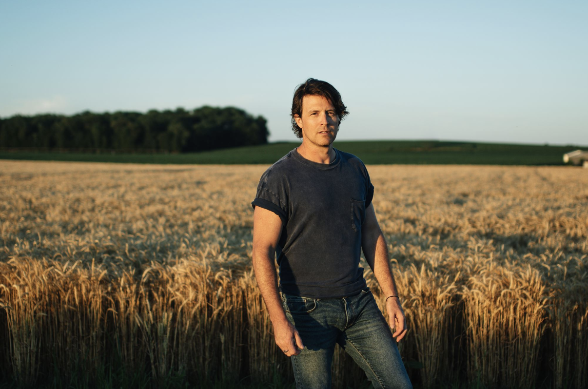A man standing in a golden wheat field against a blue sky and forested horizon.